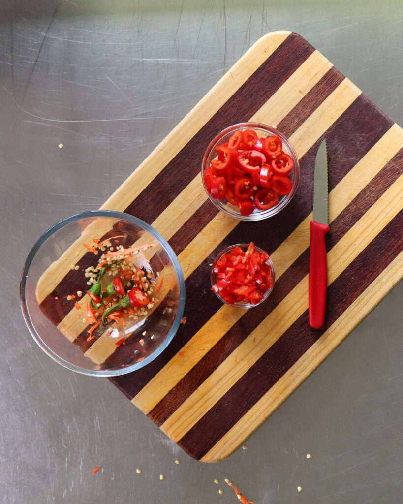 Three bowls on a striped wooden cutting board coloured with the intense red of bowls of sliced and chopped chilli. The third bowl is the discarded chilli seeds.