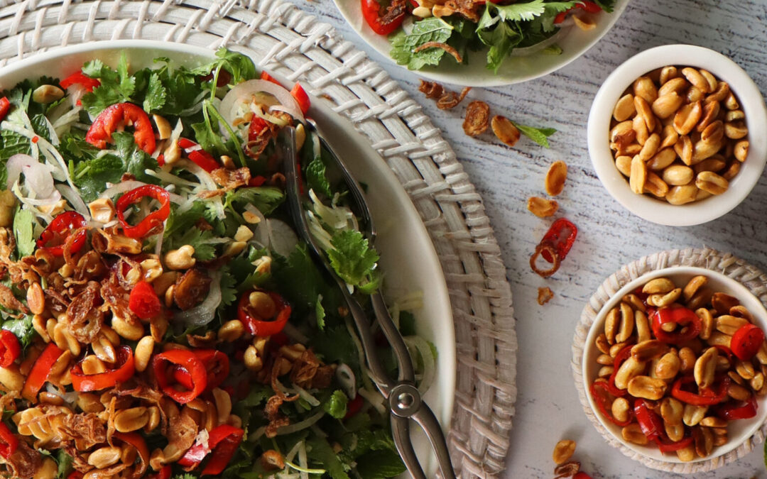 Vibrant Thai Green Pawpaw Salad in a white ceramic serving dish on a round woven placemat next to three bowls – a serving of salad, a small dish of extra peanuts, and a bowl of extra chilli mix toppings.