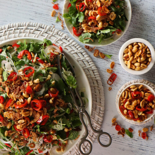 Vibrant Thai Green Pawpaw Salad in a white ceramic serving dish on a round woven placemat next to three bowls – a serving of salad, a small dish of extra peanuts, and a bowl of extra chilli mix toppings.