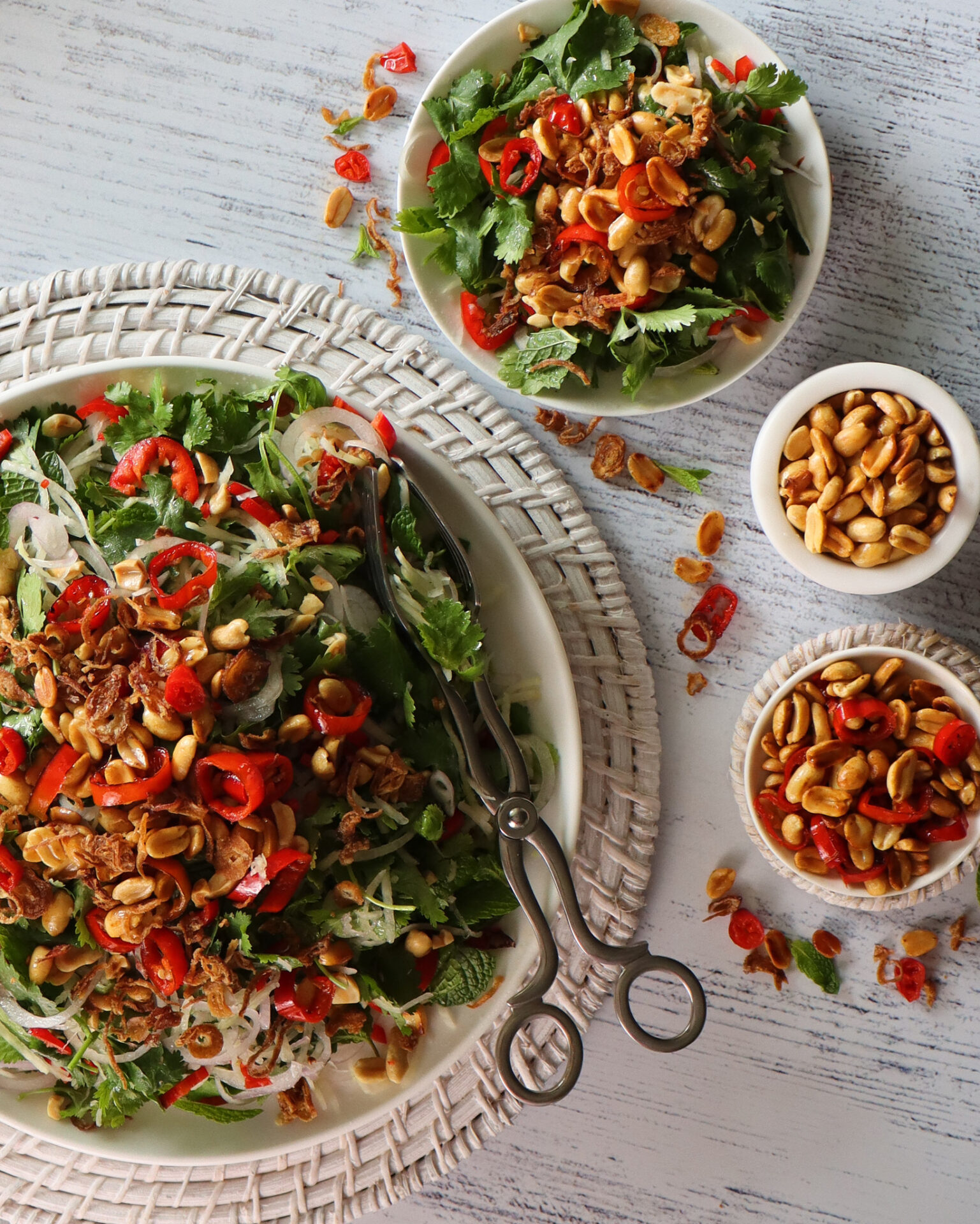 Vibrant Thai Green Pawpaw Salad in a white ceramic serving dish on a round woven placemat next to three bowls – a serving of salad, a small dish of extra peanuts, and a bowl of extra chilli mix toppings.