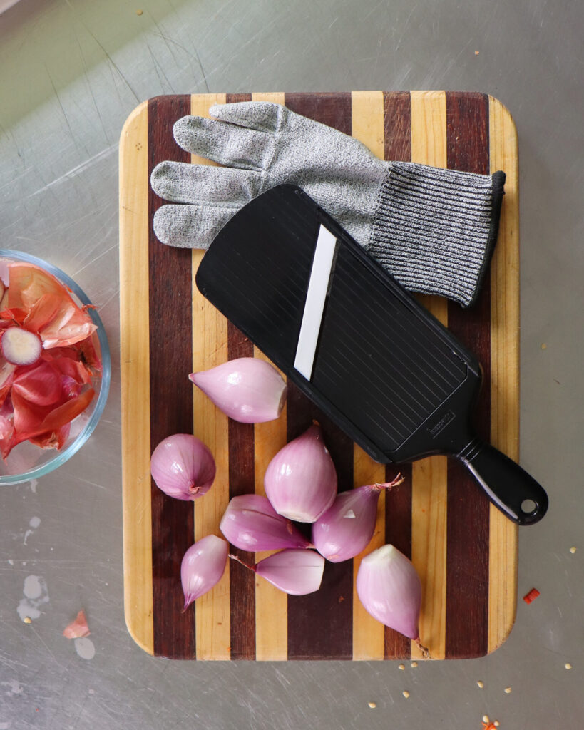 Sliced shallots next to a mandolin resting on a striped wooden cutting board next to a glass bowl of discarded shallot skins.