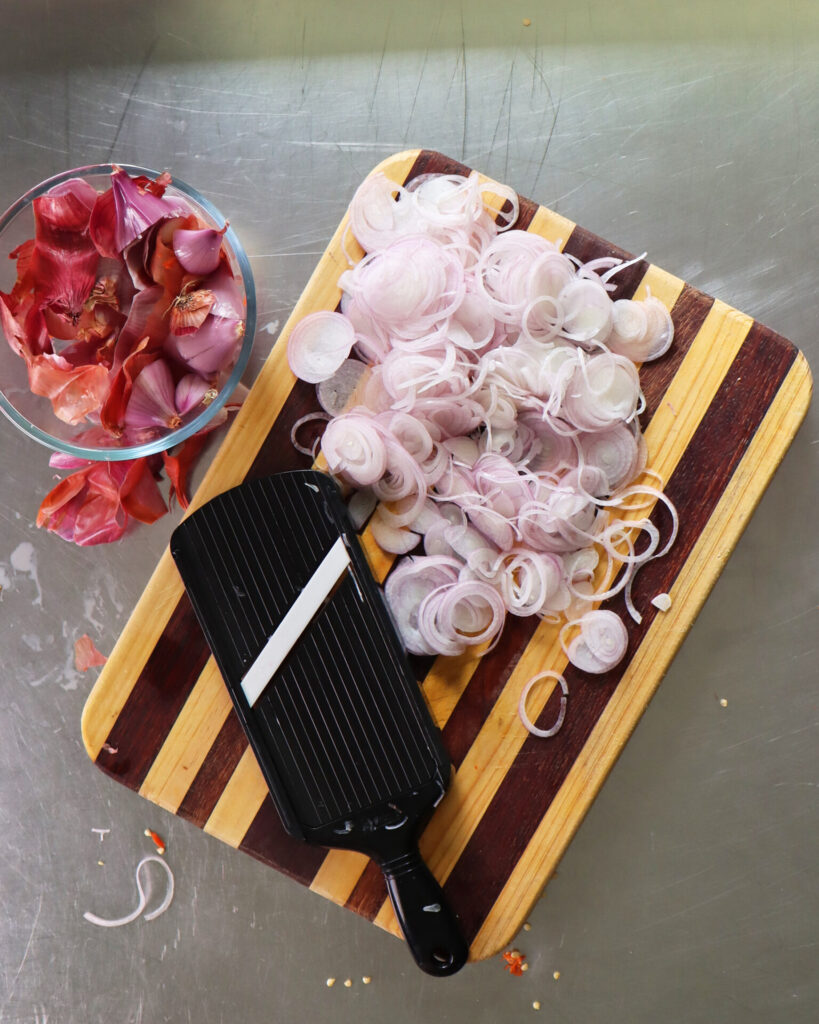 Sliced shallots next to a mandolin resting on a striped wooden cutting board next to a glass bowl of discarded shallot skins.