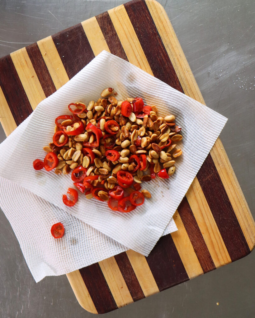 Salad garnish of peanuts, crispy shallots and chilli draining on a paper towel placed on a striped wooden cutting board.