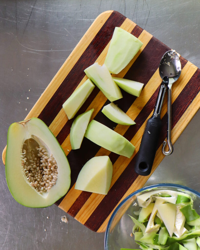 Green pawpaw cut open on a striped wooden cutting board. Half of the pawpaw has not yet been de-seeded. The other half has been peeled and roughly cut. The discarded peel is in a glass bowl next to the cutting board.