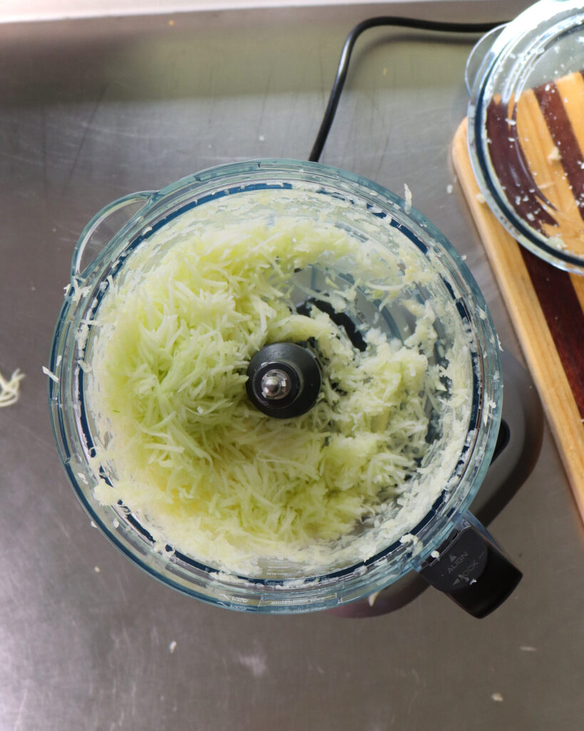 Green pawpaw flesh shredded in a food processor on a stainless-steel benchtop next to a striped wooden cutting board.