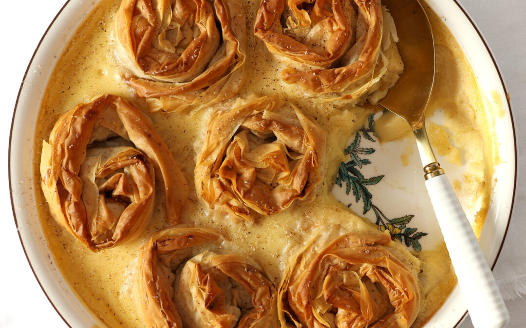 Fillo parcels filled with spiced-apple and verjuice-soaked sultanas baked in a creamy custard in a round porcelain dish on a white tablecloth.