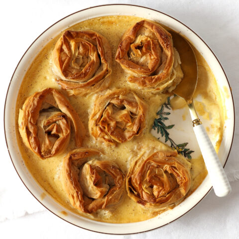 Fillo parcels filled with spiced-apple and verjuice-soaked sultanas baked in a creamy custard in a round porcelain dish on a white tablecloth.