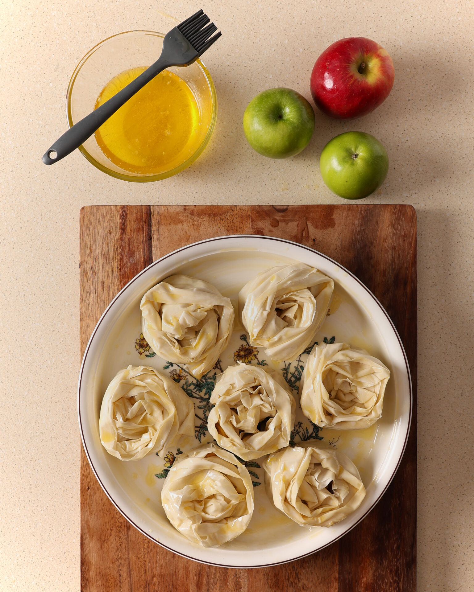 A round porcelain baking dish filled with fillo pastry rosettes for an apple and custard crinkle pie sits on a wooden board on a kitchen bench. Above the board is a bowl of melted butter and a pastry brush along with three apples.