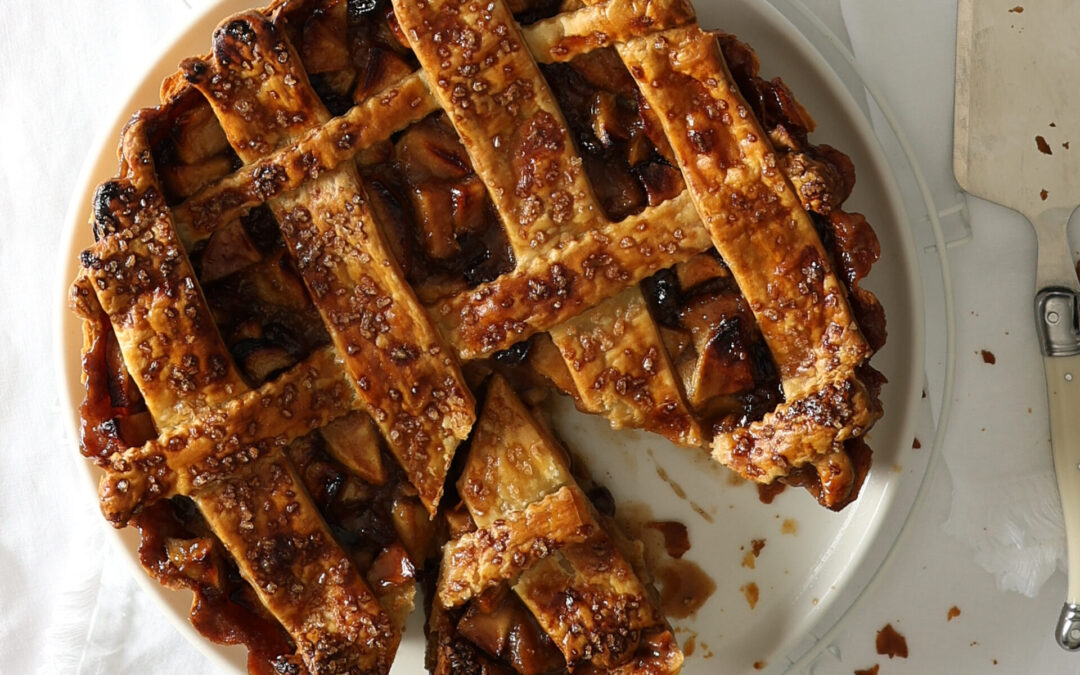 Short crust lattice apple pie with verjuice-soaked sultanas on a white tablecloth with a pie server, three silver forks and square white plates.