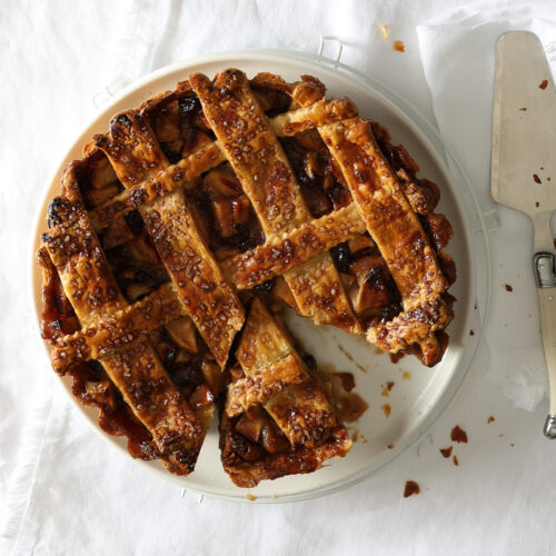 Short crust lattice apple pie with verjuice-soaked sultanas on a white tablecloth with a pie server, three silver forks and square white plates.