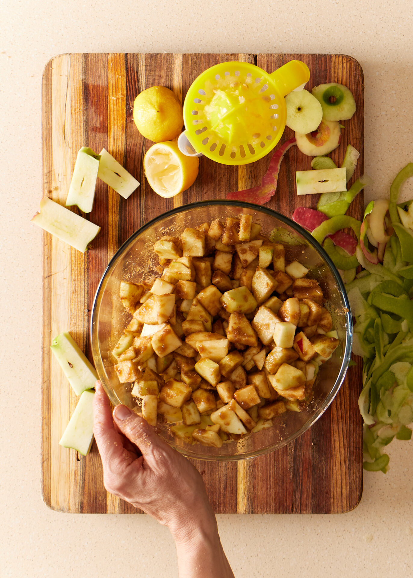 Spiced-apple pie filling in a glass bowl on a chopping board, surrounded by apple peels and lemons.