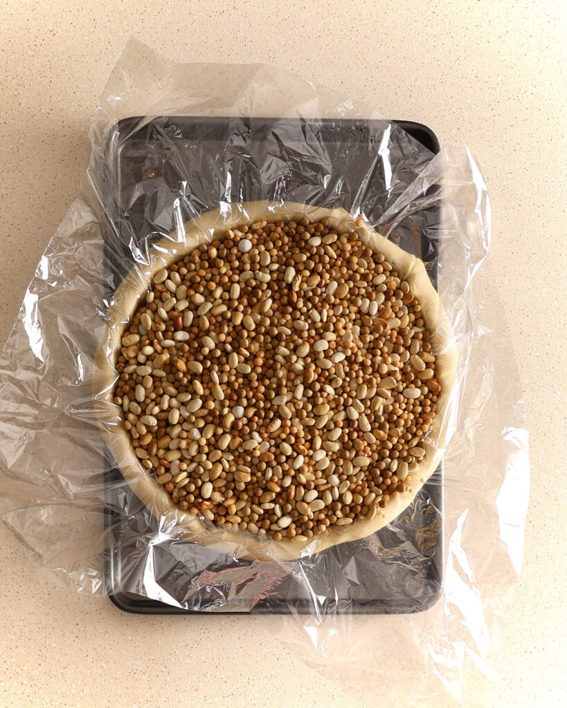 A pie pan lined with pastry , sits on a baking tray. An oven roasting bag sits over the raw pastry and underneath the dried beans that fill the pan to the top. The bottom pie crust is ready to be blind baked.