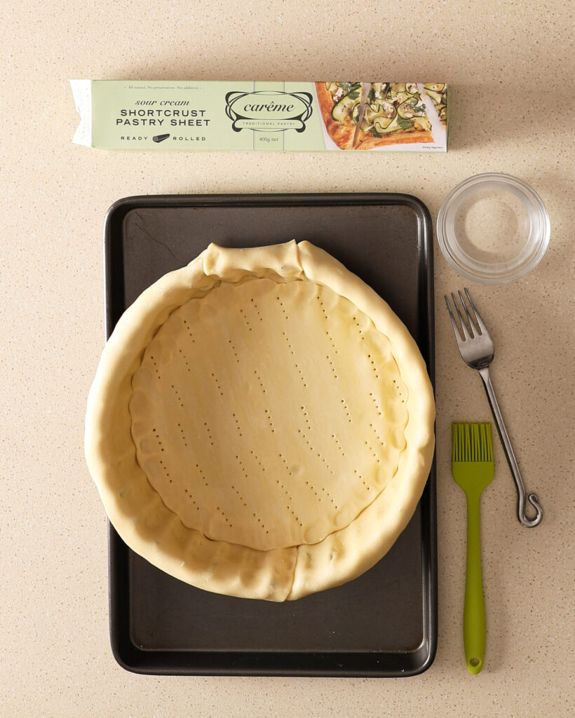 A pie pan sitting on a kitchen bench, with pastry lining the bottom of the pan. The pastry is draped over the edges of the pan. A box of Carême short crust sheet sits above the pie pan.