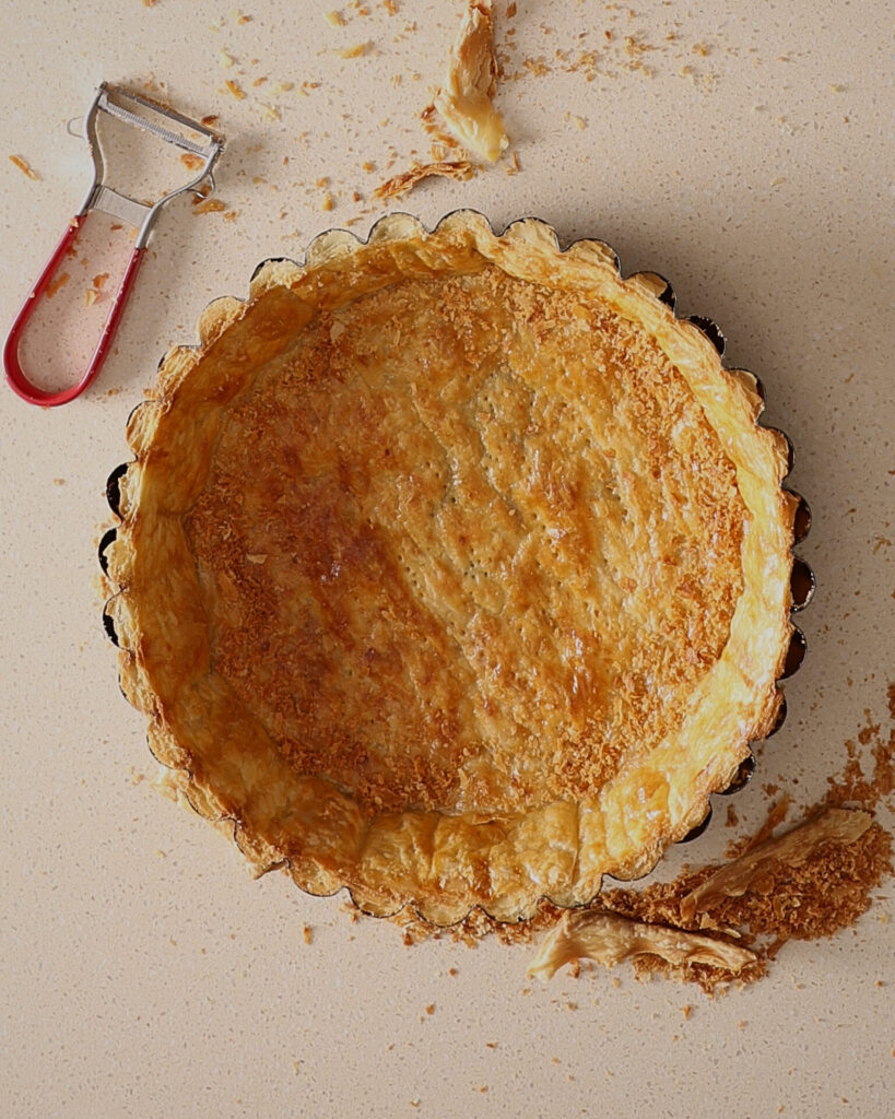 A golden blind baked pie crust sits on a kitchen bench in a fluted pie pan. There is a swivel style vegetable peeler on the bench. The top edge of the crust has been trimmed back to reveal the fluted sides of the pan. Pastry crumbs surround the pie crust.