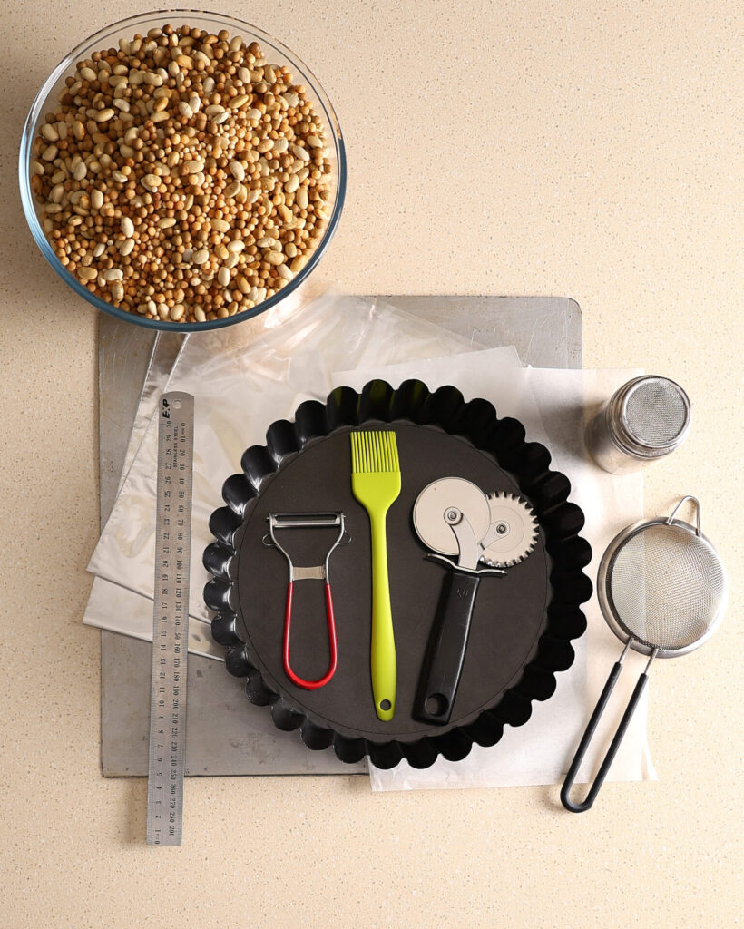 A collection of equipment needed to make the apple pie pastry crusts. On a kitchen bench is a bowl full of dried beans, fluted pie pan, baking tray, ruler, swivel grater, pastry brush, pastry wheel, small sieve, baking paper, oven roasting bag, and flour shaker.