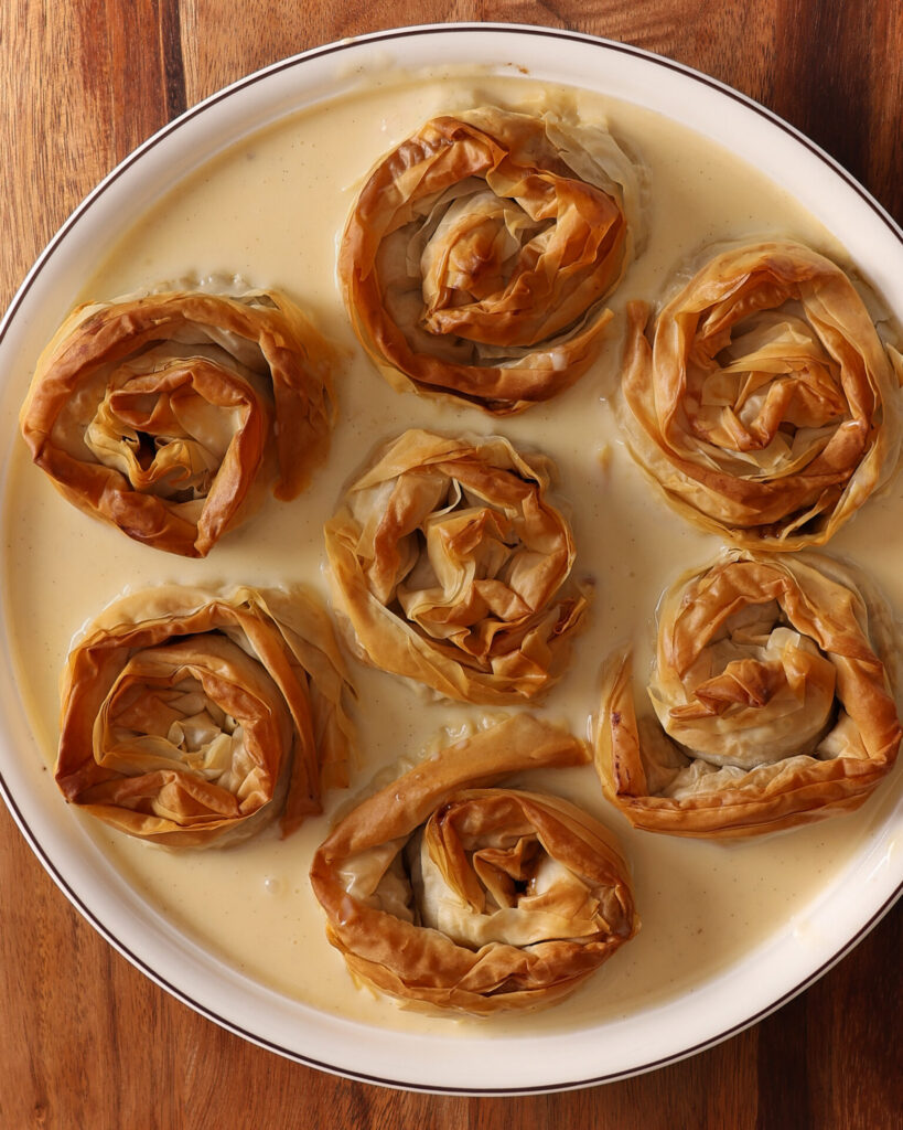 A round porcelain baking dish sits on a wooden board. The dish full of seven baked fillo pastry rosettes surrounded by just poured vanilla custard. This dish is about to be returned to the oven to bake the custard for this apple and custard crinkle pie.