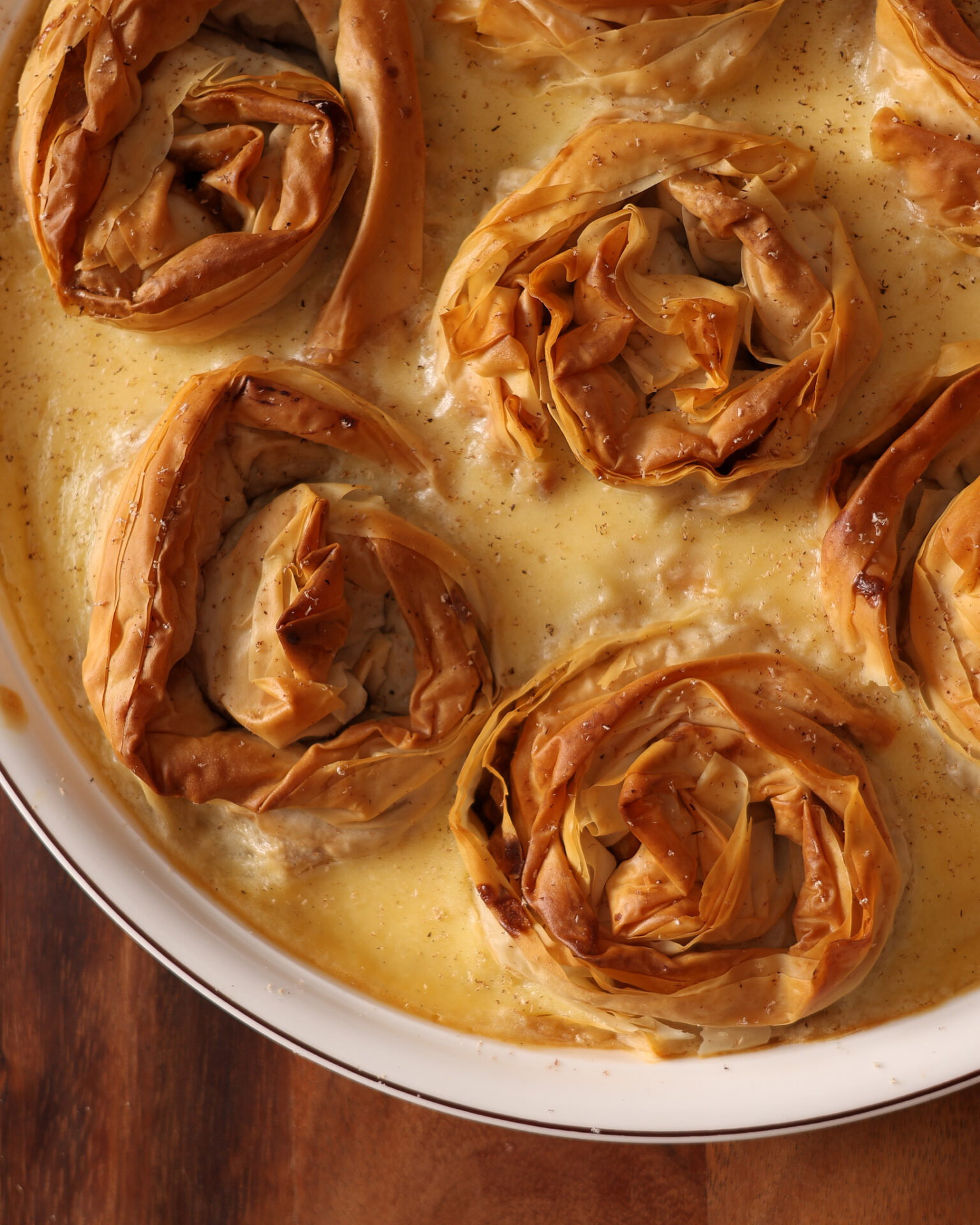 Close up of a spiced-apple and custard crinkle pie made with apple filled fillo rosettes surrounded by baked vanilla custard. The pie was baked in a round porcelain dish that sits on a wooden board.