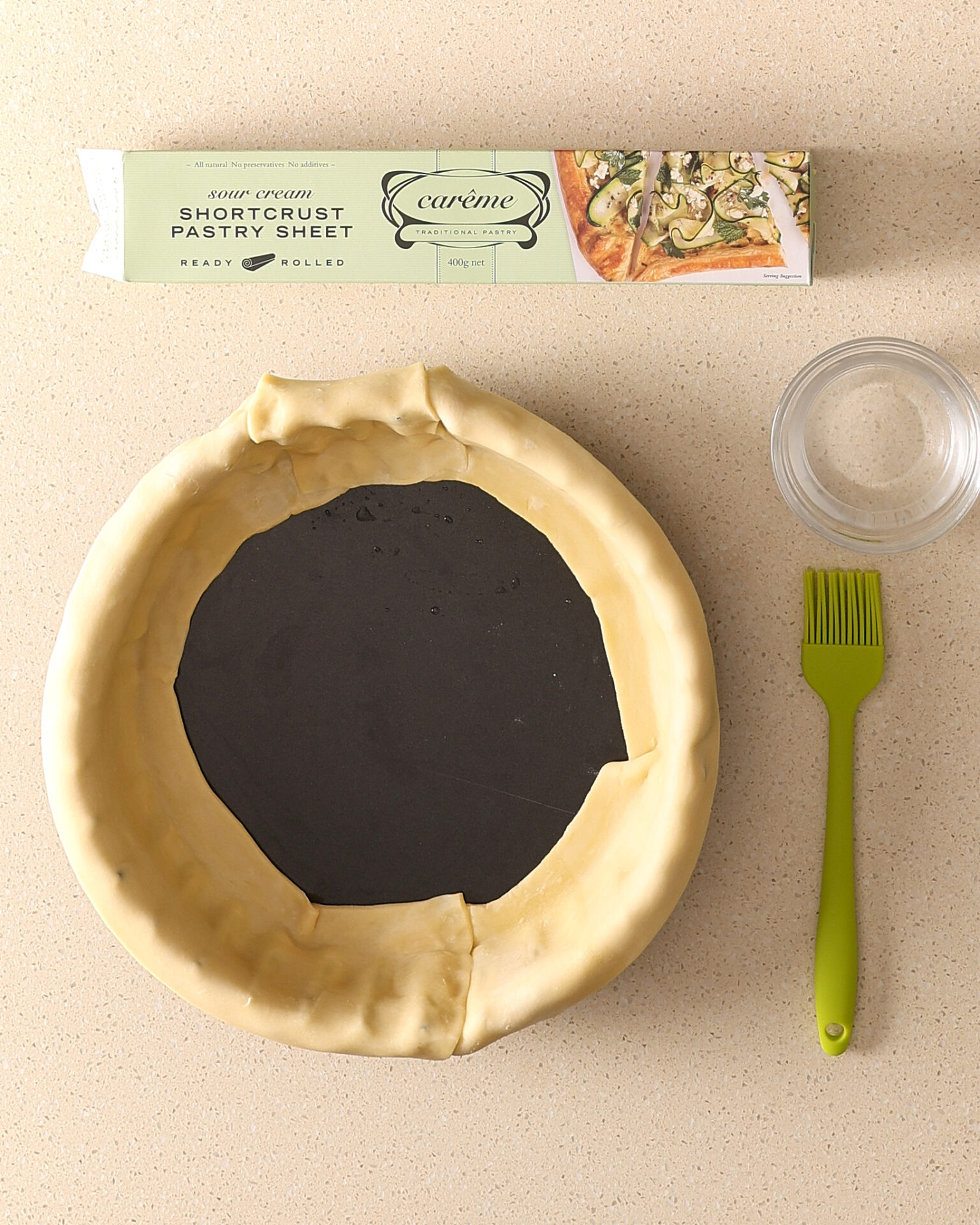 A fluted pie pan is sitting on a kitchen bench, strips of short crust pastry lines the sides of the pan and the bottom of the pie shell is not yet in place. A box of Carême short crust pastry sits above the pie pan.