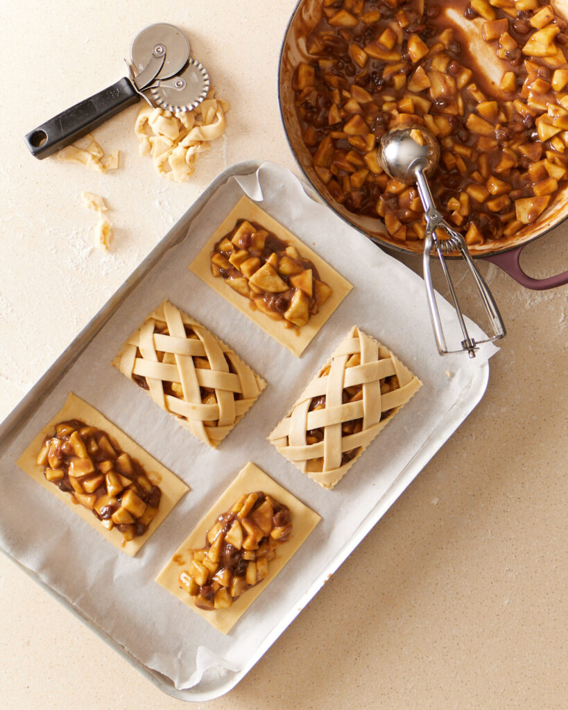 Apple hand pies being assembled on a kitchen bench. The apple and raisin pie filling is in a casserole dish. Five partly assembled rectangular pies sit on a baking paper lined tray. Two of the pies are finished with a lattice top crust.