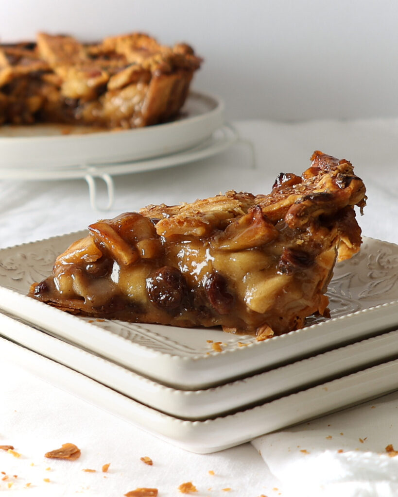 Slice of pie on a stack of three square ceramic plates on a white table cloth sprinkled with flaky crust. The lattice pie is on a rack in the background.