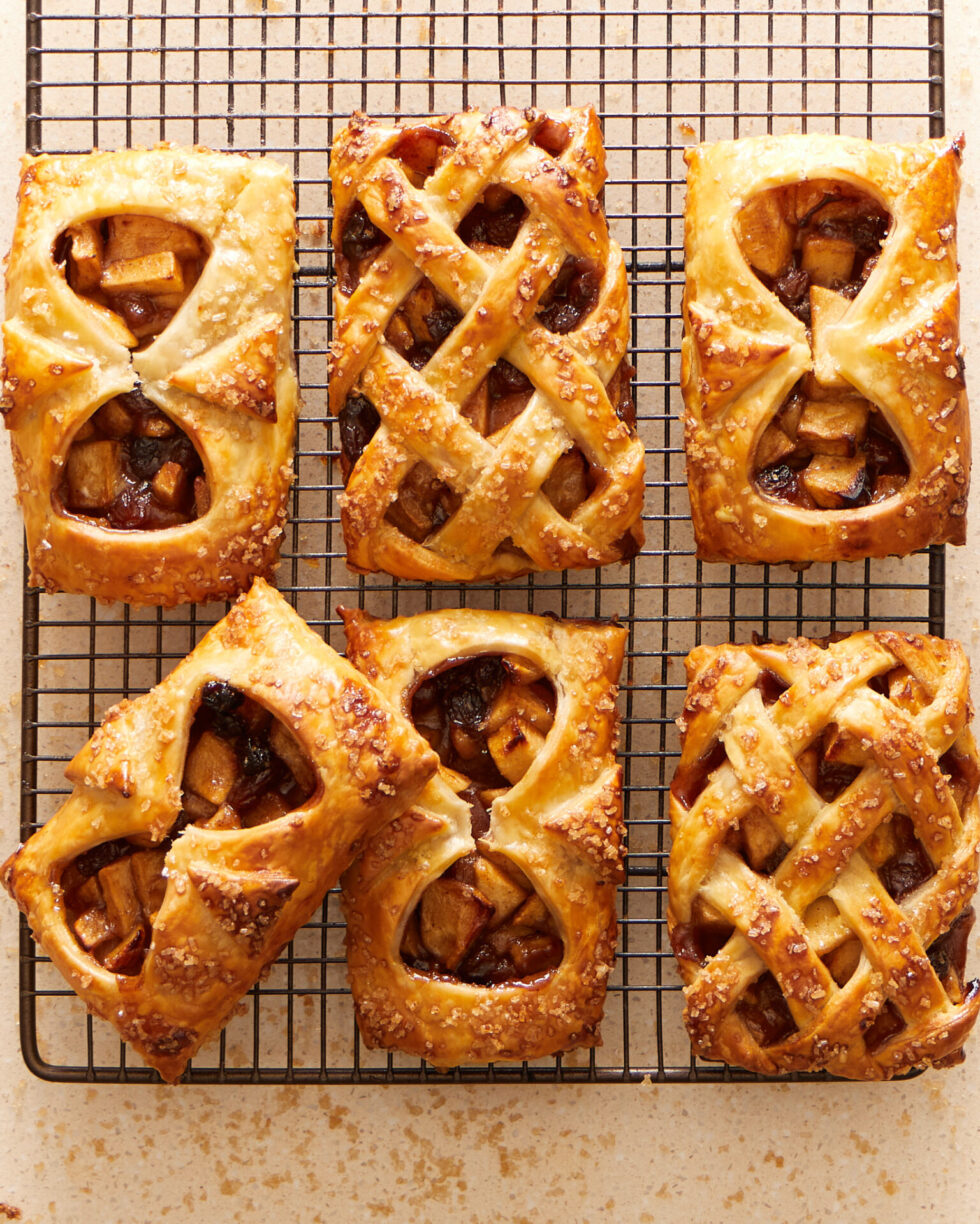 Six baked spiced-apple and sultana danishes made with Carême short crust pastry sit on a cooling rack. Two have lattice crusts and four crusts with cut outs showing the apple filling inside.