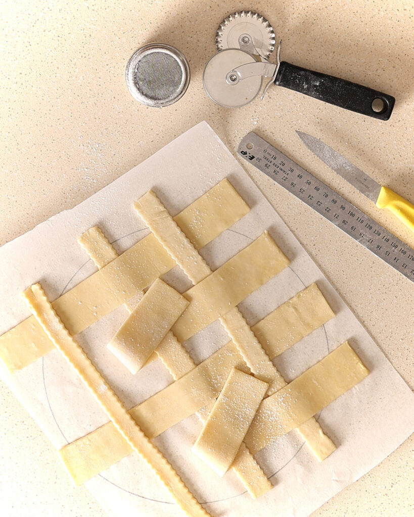 Pastry on a kitchen bench being assembled into a lattice crust on baking paper. The equipment used is on the bench and includes a ruler, pastry reel, paring knife, and flour shaker.