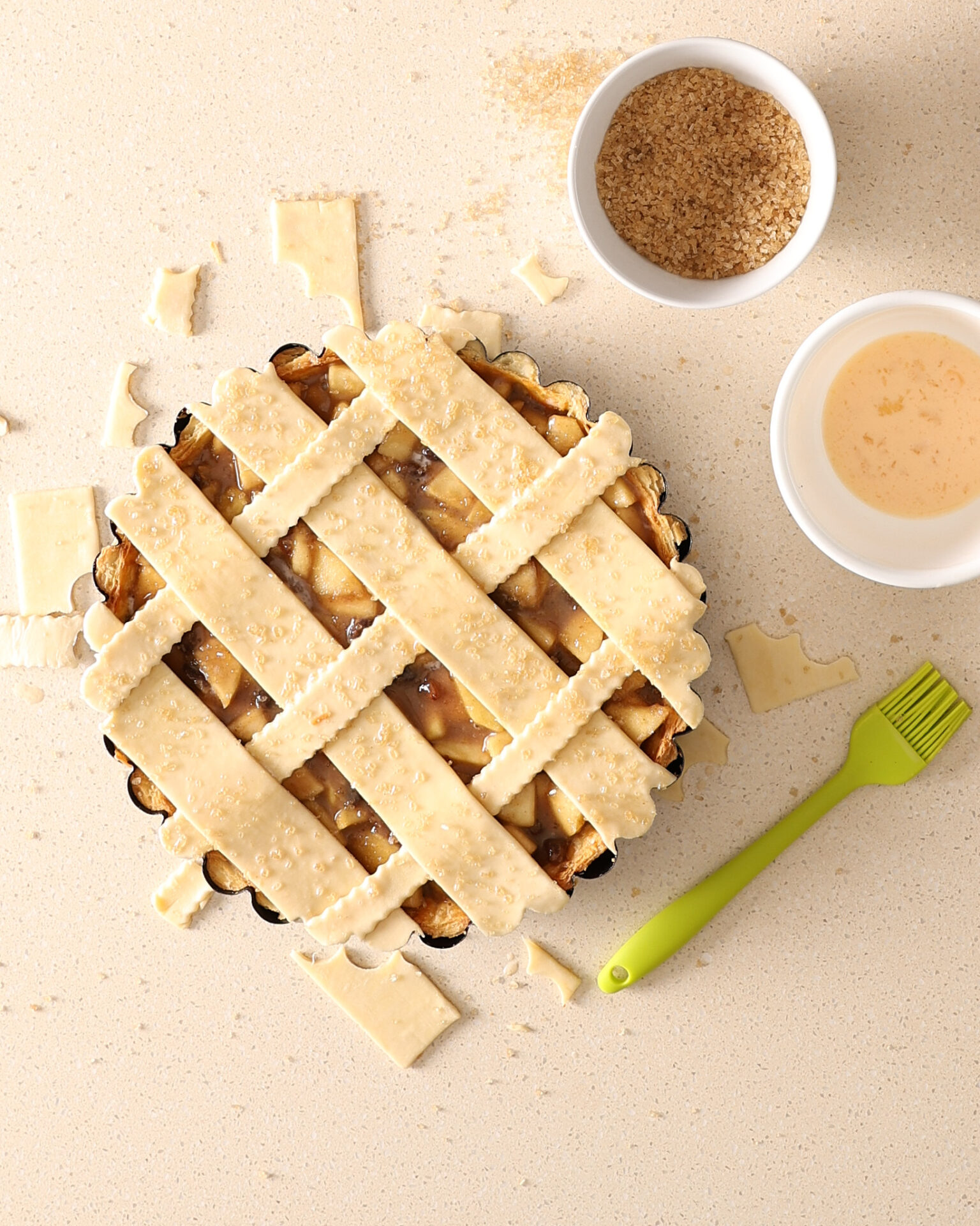 Assembled short crust lattice apple pie on a kitchen bench. Egg wash, a bowl of coffee sugar and a pastry brush sit beside the pie. Pastry scraps lie on the bench around the spiced-apple pie.