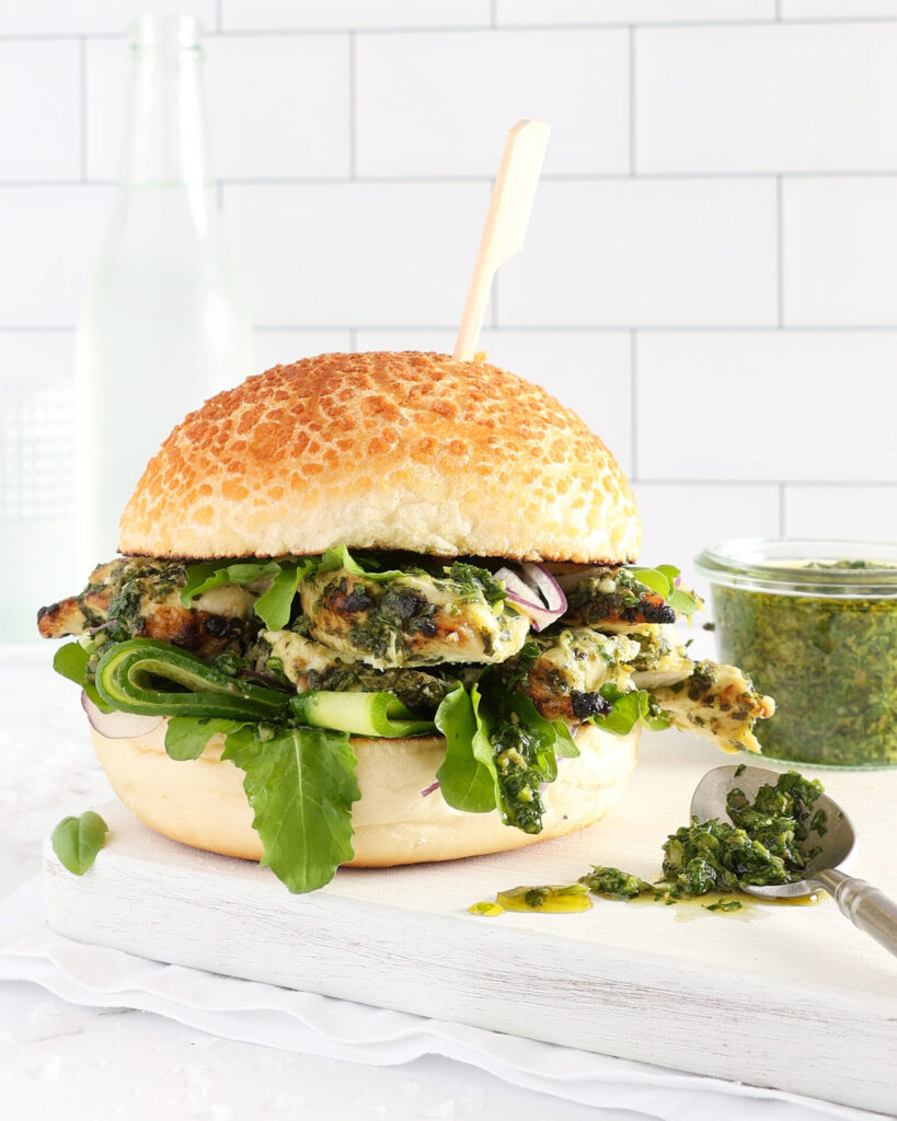 A chicken burger is sitting on a white wooden board and white cloth napkin. The burger is made from salsa verde mayonnaise chicken breast kebab slices, sliced cucumber, red onion, and rocket. A small glass jar of salsa verde sits beside the burger and a metal spoon sits with some spilled sauce beside the burger. The burger bun is held in place with a wooden skewer. Clear glass bottles are in the background against a white tiled wall.