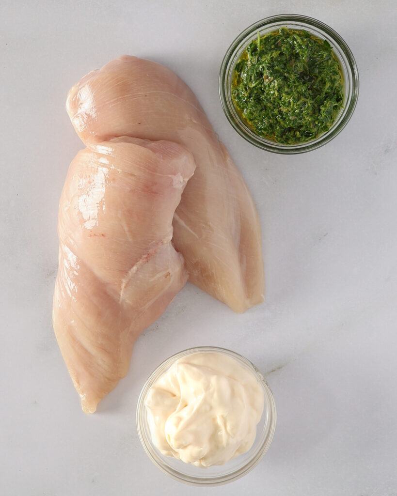 Two skinless chicken breasts sit on a marble bench. There is a small bowl of Italian salsa verde above the chicken breasts and a small bowl of mayonnaise below. These are the ingredients for a salsa verde chicken recipe.