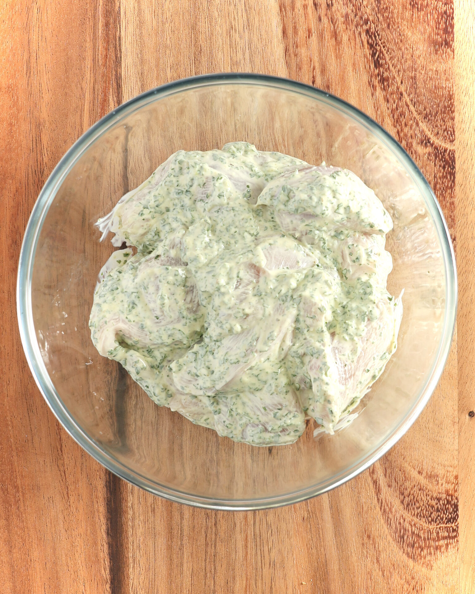 Sliced chicken breasts are marinating in a glass bowl on a wooden board. The marinade is made from mayonnaise an Italian herb sauce as part of a salsa verde mayonnaise chicken breast kebab recipe.