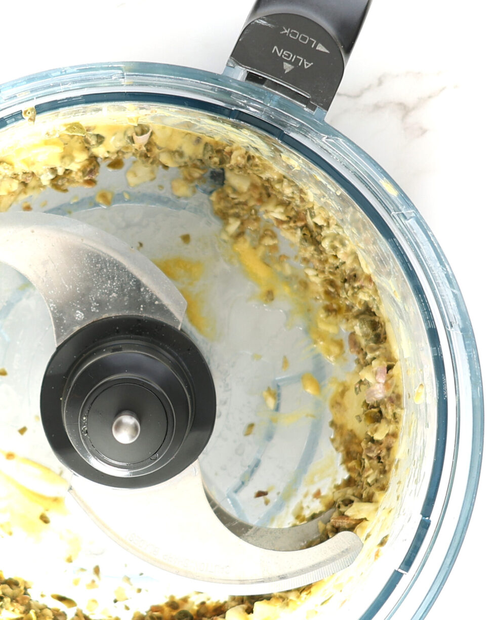 A close-up of the food processor bowl with a chopping blade. Capers, garlic, and anchovies are finely chopped with Dijon mustard and white sugar.