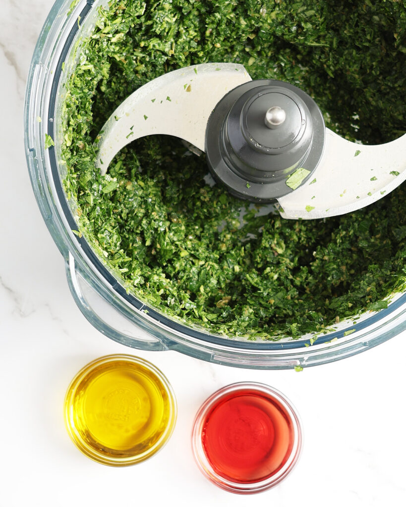 A close-up of salsa verde in the food processor bowl with the chopping blade. The basil and flat parsley are finely chopped in the bowl. Olive oil and red wine vinegar sit in mini glass bowls beside the food processor.