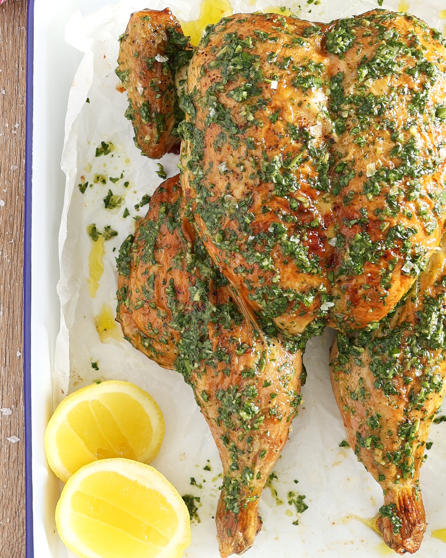 A close-up of a roast butterflied chicken on baking paper in a white metal roasting pan. The chicken has been brushed with salsa verde Italian green sauce. A halved lemon sits below the chicken.