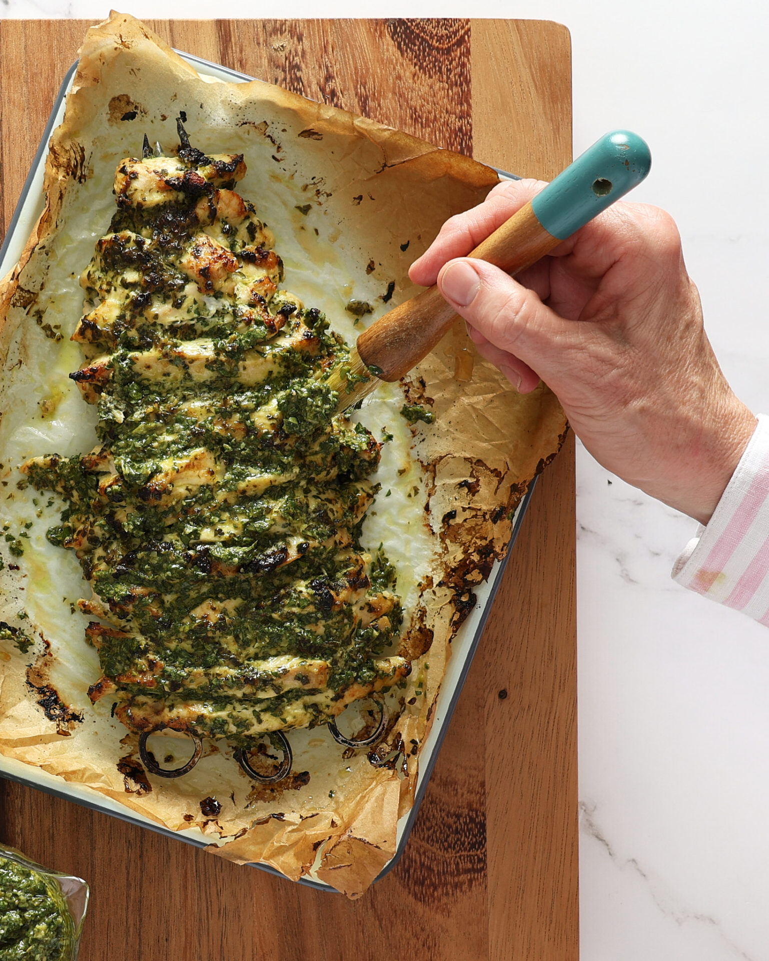 A salsa verde mayonnaise chicken breast kebab is sitting on baking paper in a white enamel roasting pan sitting on a wooden board on a marble benchtop. It has just been taken out of the oven and is golden brown with green herbs. A hand is seen holding a pastry brush that is brushing more Italian salsa verde over the kebab.