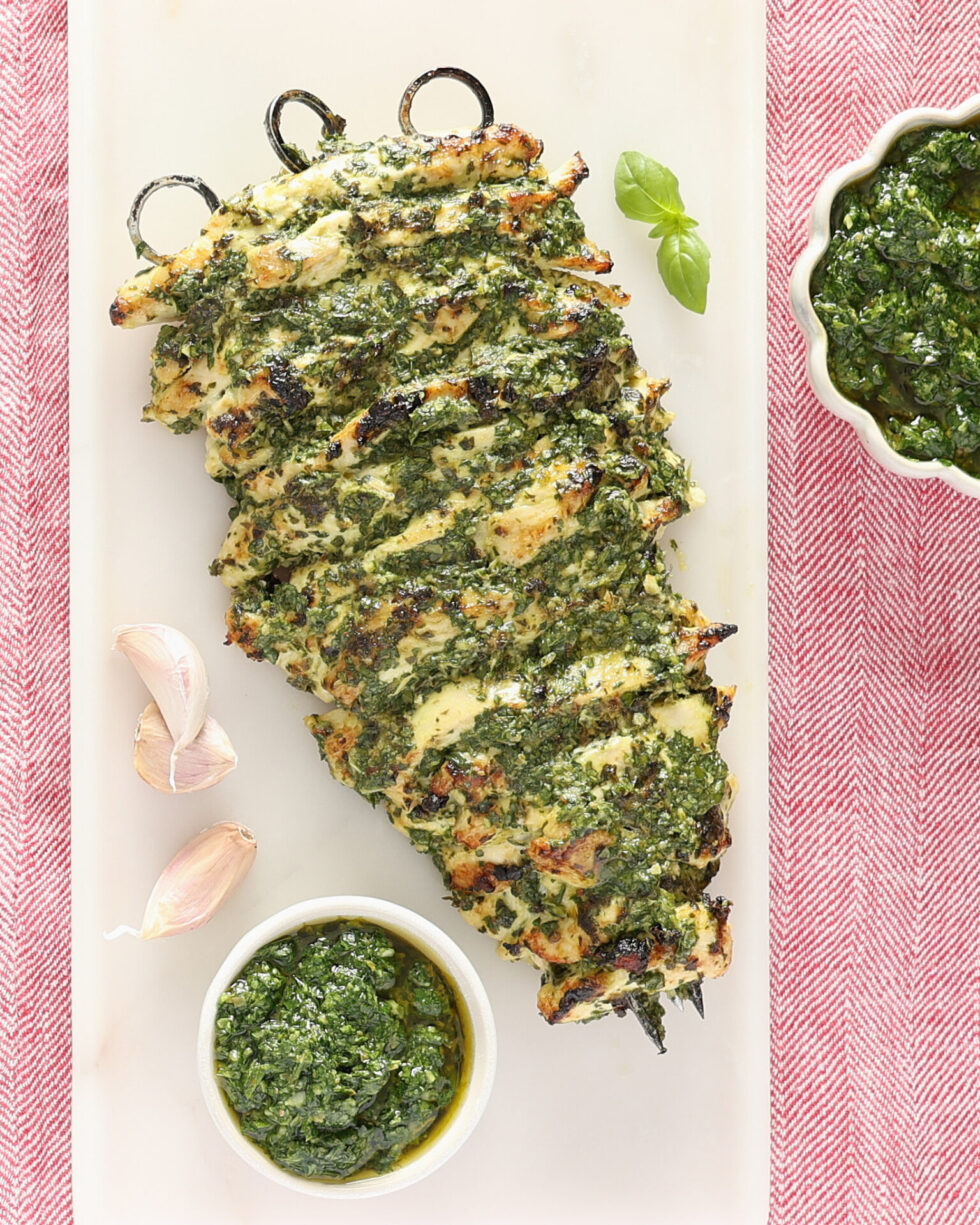 A cooked salsa verde mayonnaise chicken breast kebab sits on a rectangular marble plate and red cloth. The kebab is made with strips of chicken that have been skewered onto 3 metal skewers. The kebab has been basted with salsa verde there are two small bowls of salsa verde, one above the kebab and one below. Three garlic cloves are on the plate beside the kebab.
