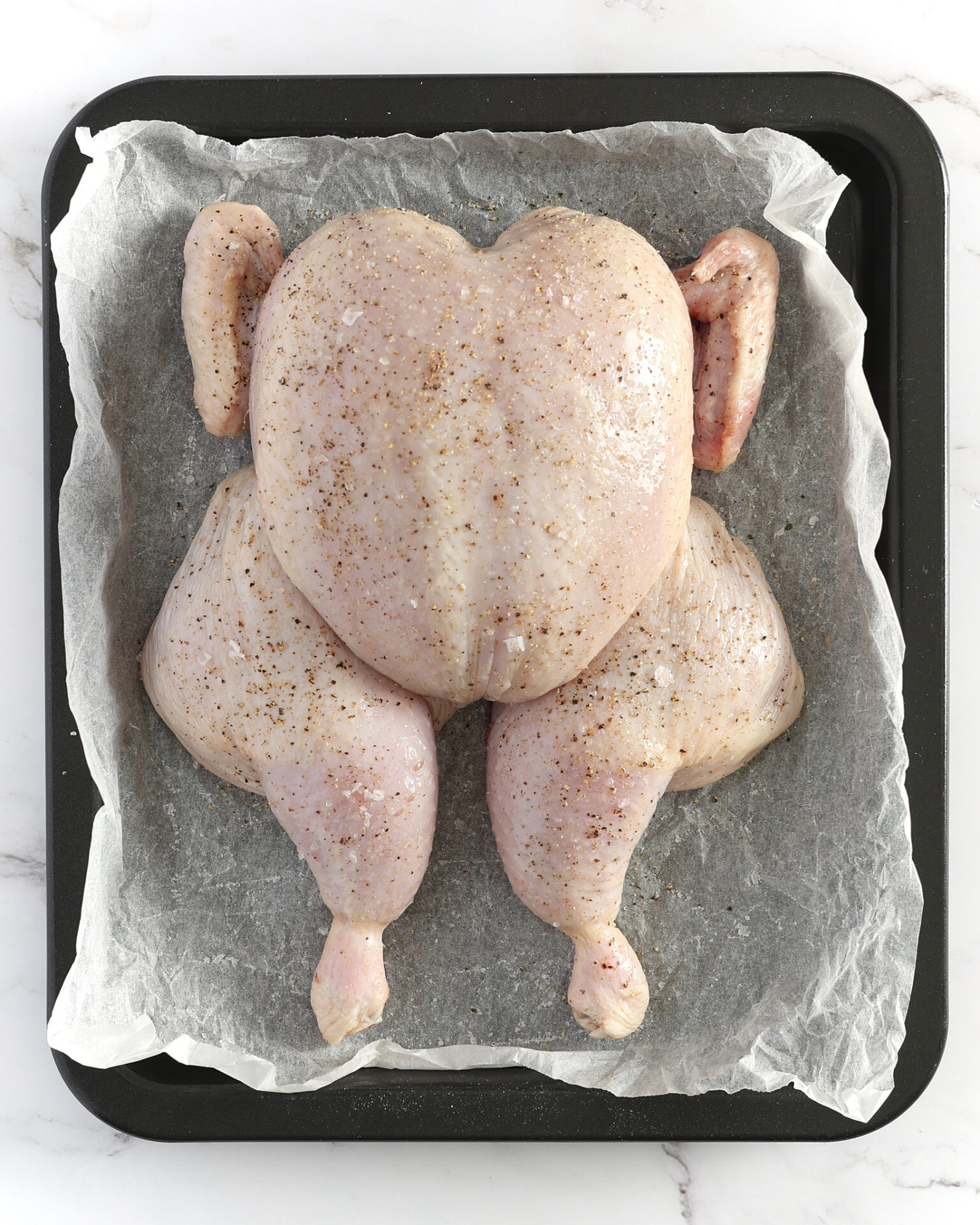 As part of a spatchcocked roast chicken recipe, a raw butterflied chicken sits on baking paper in a black roasting pan. The chicken is ready for the oven and has been seasoned with salt and pepper.