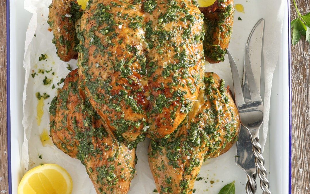 A roast spatchcocked chicken sits on a wooden bench in a white metal roasting pan. The chicken has been brushed with salsa verde Italian green sauce and a bowl of the sauce sits above the chicken. There is a red cloth at the top of the image. Halved lemons sit beside the chicken and a large carving knife and fork are placed on the opposite side.