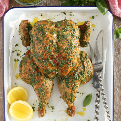 A roast spatchcocked chicken sits on a wooden bench in a white metal roasting pan. The chicken has been brushed with salsa verde Italian green sauce and a bowl of the sauce sits above the chicken. There is a red cloth at the top of the image. Halved lemons sit beside the chicken and a large carving knife and fork are placed on the opposite side.