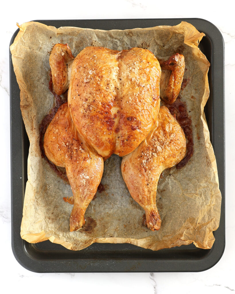 A roasted butterflied whole chicken on baking paper in a shallow roasting pan. The chicken is straight out of the oven and seasoned with salt and pepper.