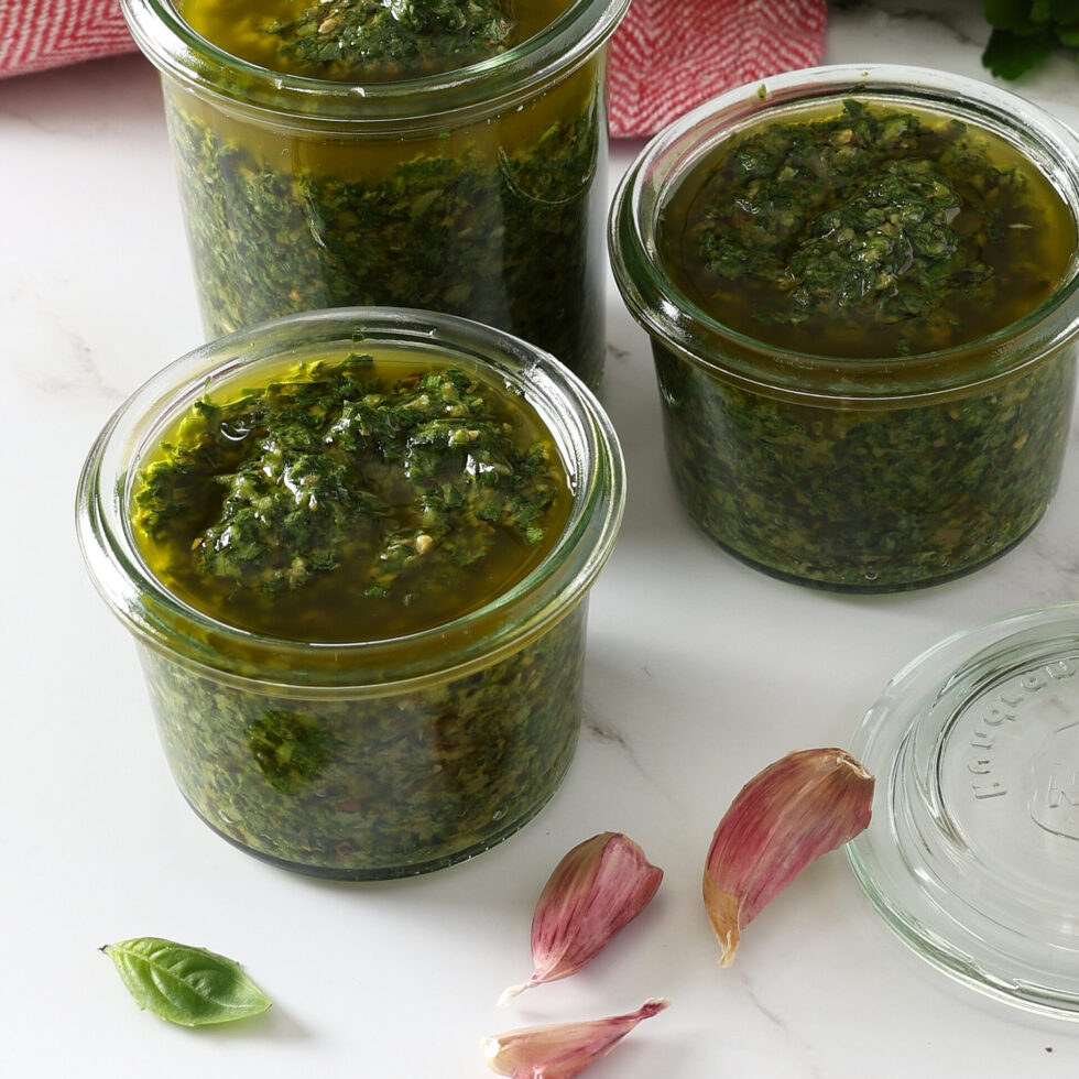 Vibrant green salsa verde sauce in three small jars on a marble bench with three cloves of garlic in front of the jars.