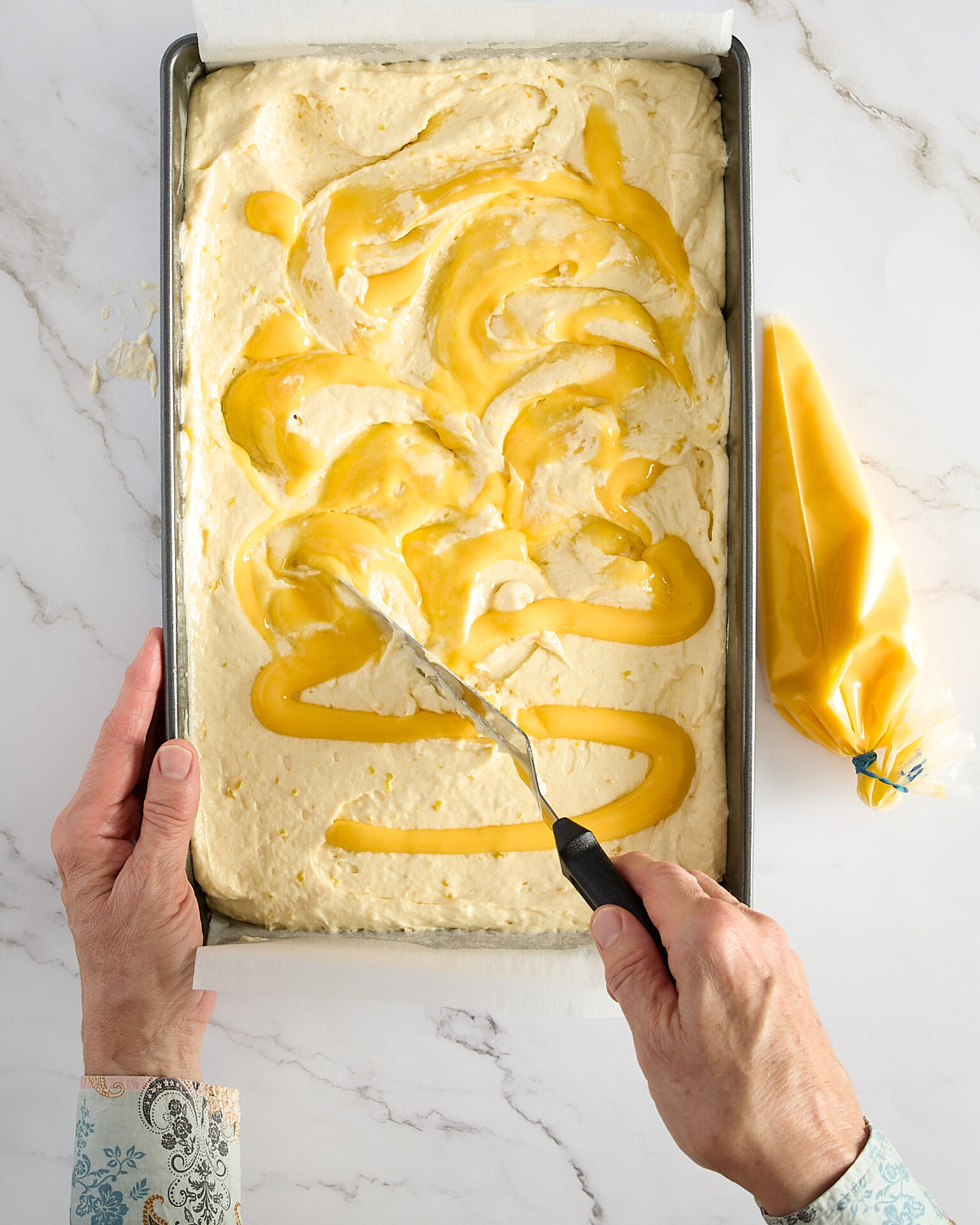 Cook is swirling the lemon curd through the cake batter in a sheet cake pan, a piping bag sits to the side.