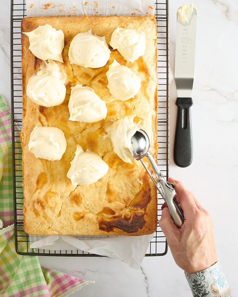 Cook is placing scoops of frosting over the top of the cake on its cooling rake.