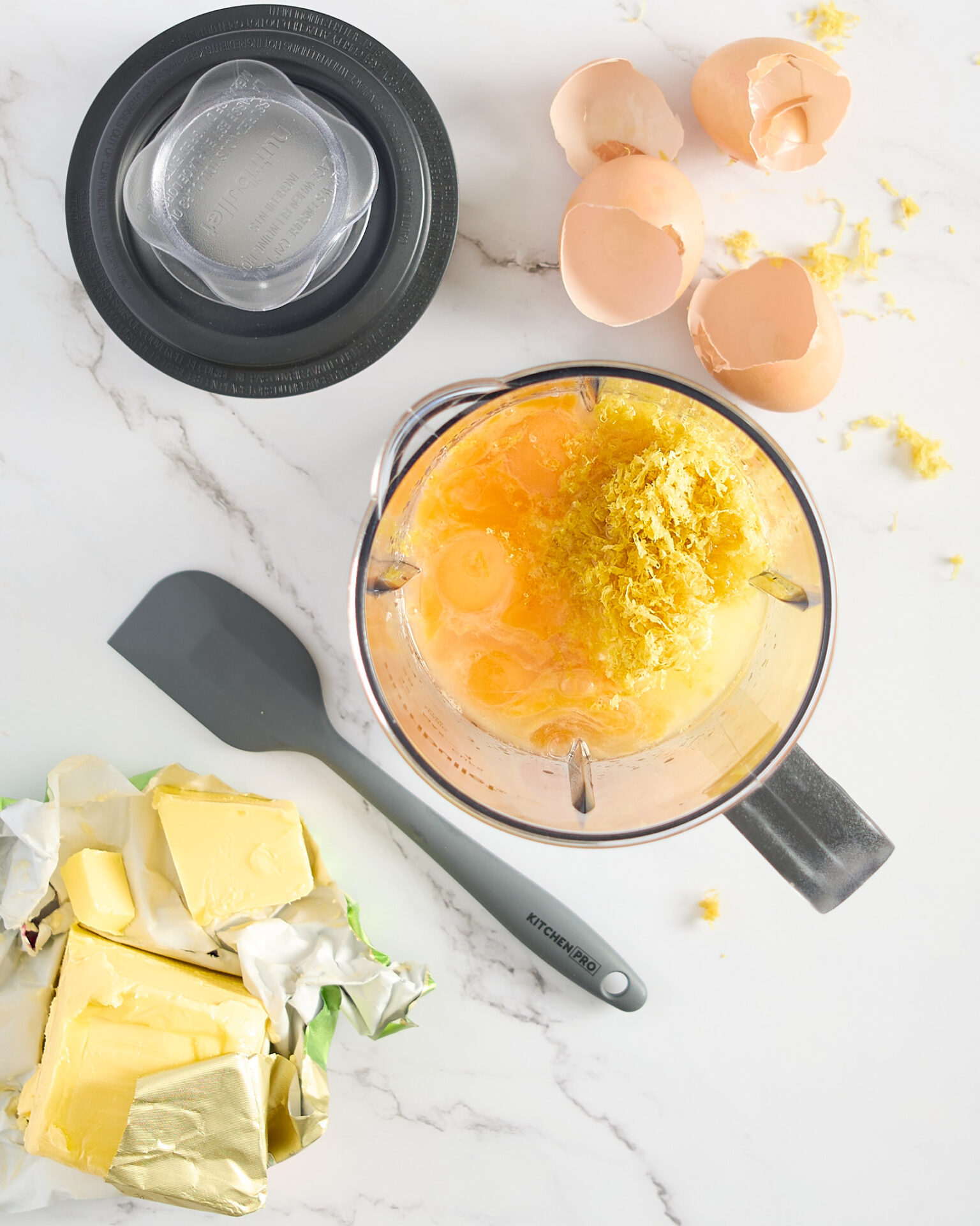 Lemon curd ingredients in the jug waiting to be blended sitting on a marble benchtop with a few ingredients to the side.