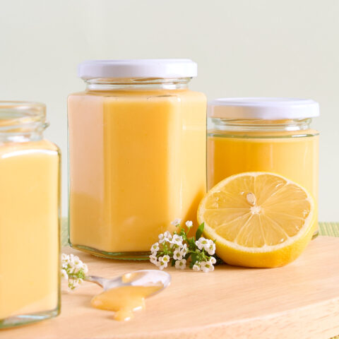 Lemon curd jar sit on a wooden board with lemon blossoms and half a lemon.