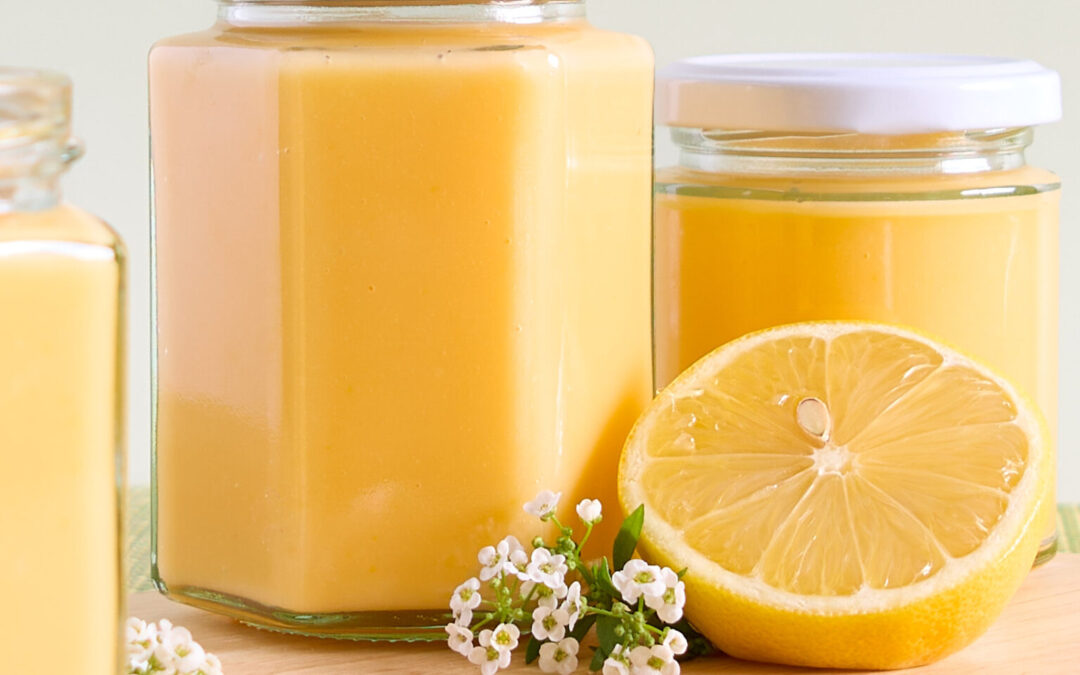 Lemon curd jar sit on a wooden board with lemon blossoms and half a lemon.