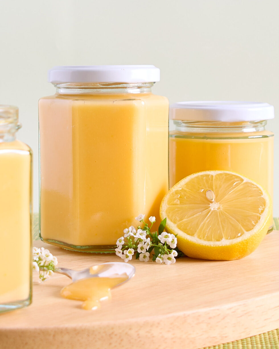 Lemon curd jar sit on a wooden board with lemon blossoms and half a lemon.