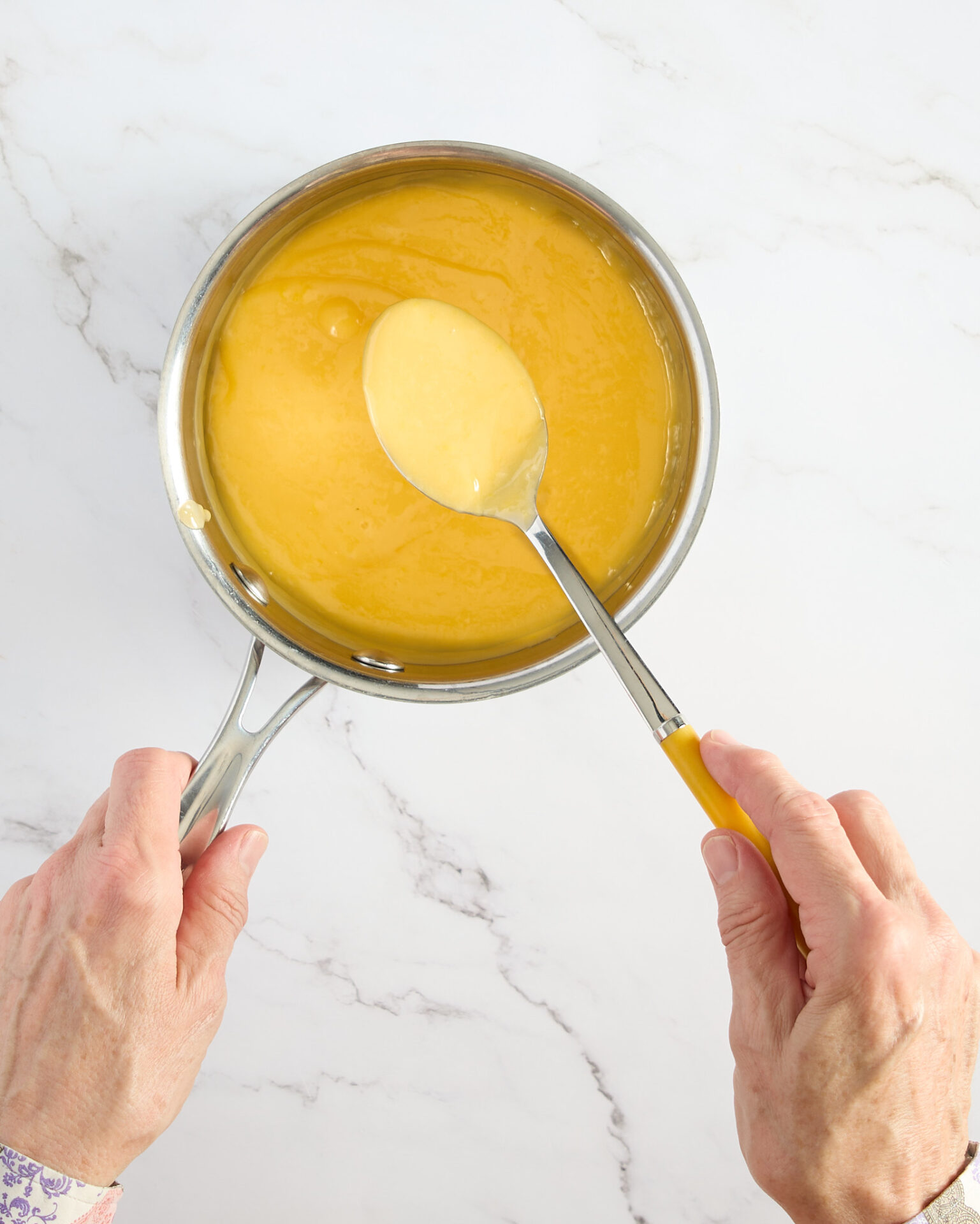 Cook shows a spoon of cooked lemon curd over the heavy bottomed stainless-steel pan.