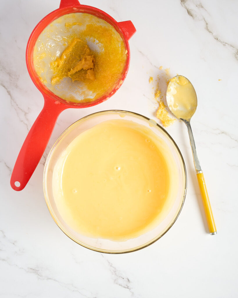 Lemon curd strained in a glass bowl with the removed zest to the side in a sieve.