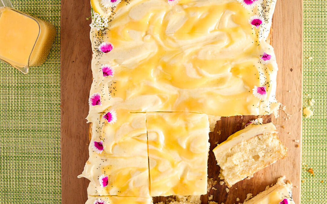 Frosted lemon sheet cake decorated with edible flowers, cut and ready to serve with fresh lemons and extra curd sitting to the side.