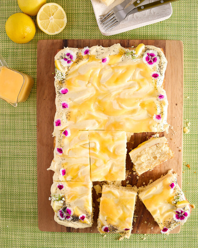 Frosted lemon sheet cake decorated with edible flowers, cut and ready to serve with fresh lemons and extra curd sitting to the side.